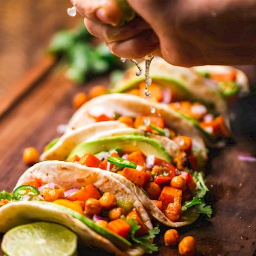 A hand squeezing lime over a row of tacos filled with chickpeas, diced vegetables, and avocado; garnished with cilantro on a wooden board.