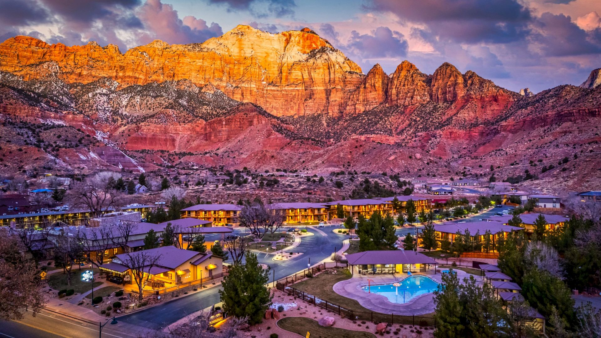 A scenic resort at sunset with illuminated buildings and a pool, set against dramatic, colorful mountains and a vibrant sky in the background.