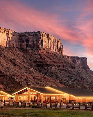 A ranch-style building with glowing lights sits at the base of a rocky cliff under a colorful sunset sky.