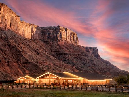 A ranch-style building with glowing lights sits at the base of a rocky cliff under a colorful sunset sky.
