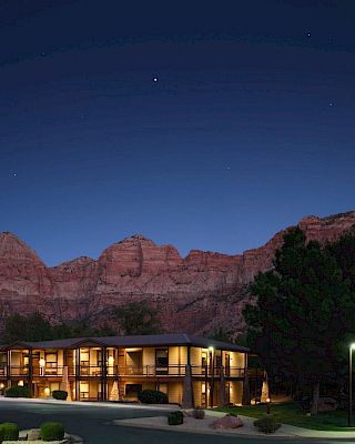 A building is illuminated at night, surrounded by trees, against a backdrop of mountains and a starry sky.