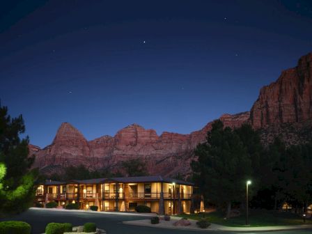 A building is illuminated at night, surrounded by trees, against a backdrop of mountains and a starry sky.