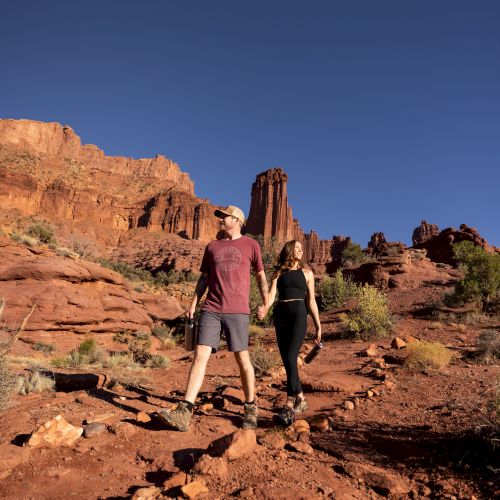 Two hikers, a man and a woman, walk along a rocky red canyon trail under a clear blue sky, surrounded by cliffs and desert scrub.