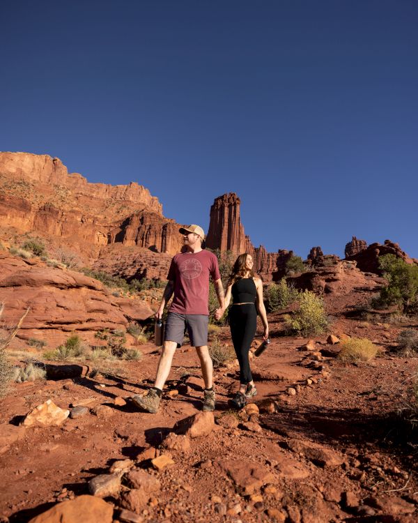 Two hikers, a man and a woman, walk along a rocky red canyon trail under a clear blue sky, surrounded by cliffs and desert scrub.