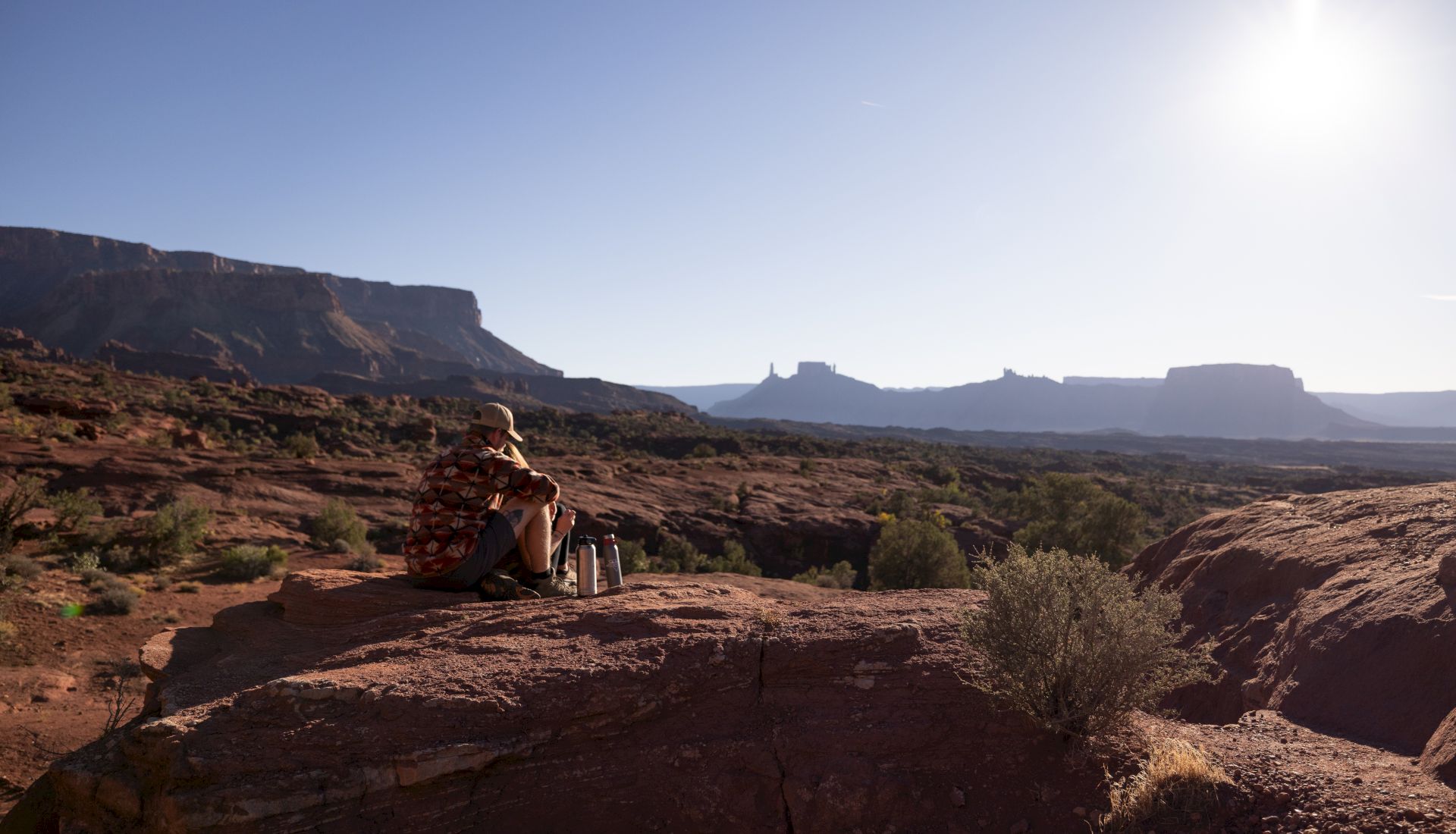 A hiker sits on a rocky overlook in a desert canyon, backpack and water bottles nearby, with mountains and mesas in view.