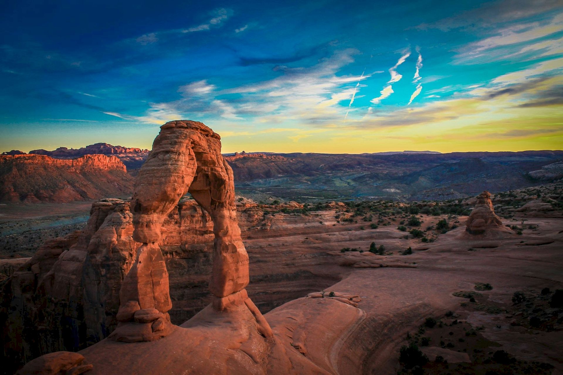 A natural rock arch formation in a desert landscape at sunset, with vibrant skies and distant mountains.