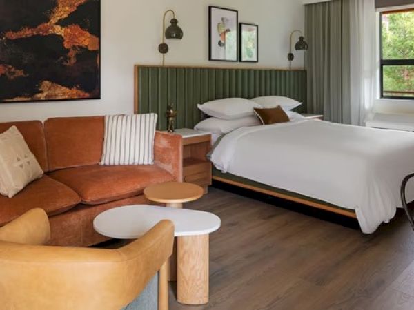 A cozy hotel room with a bed, brown sofa, armchair, and side tables, featuring artwork and a large window with greenery outside.