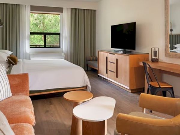 A cozy hotel room with a bed, sofa, TV, desk, chairs, and window drapes in a neutral color palette, creating a warm and inviting space.