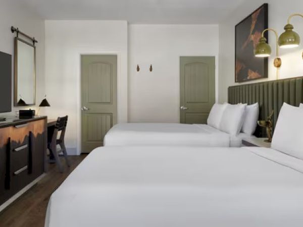 A hotel room with two beds, a TV, a desk with a lamp, two green doors, and wall art. The space appears modern and well-lit.