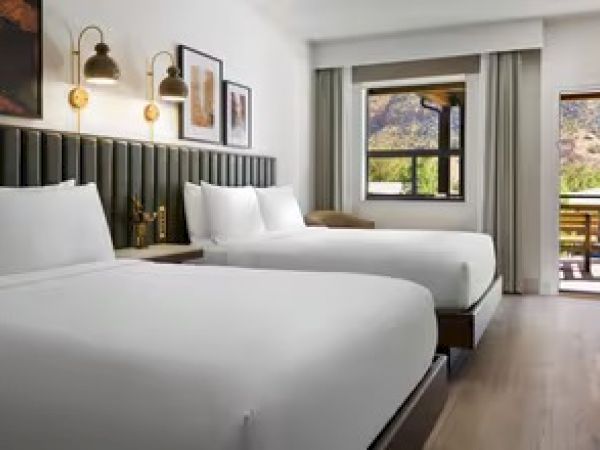 The image shows a hotel room with two beds, stylish lighting, wall art, and a view of the mountains through the window and balcony.