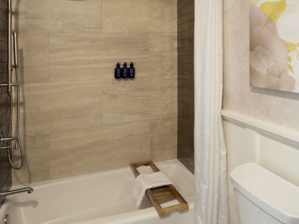 A modern bathroom featuring a bathtub with a wooden tray, shower fixtures, wall tiles, a toilet, and a piece of floral artwork.