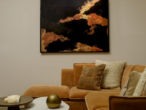 A cozy living room with a brown sectional sofa, abstract artwork on the wall, and two small tables, one with a spherical decor, enhance the space.