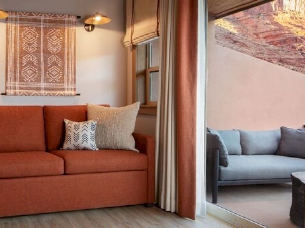 The image shows a cozy room with an orange sofa, decorative pillows, wall art, and a window view of an outdoor seating area with a scenic backdrop.