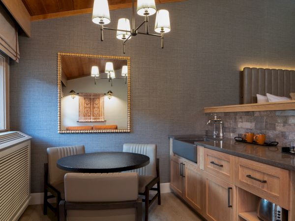 A cozy kitchenette with a small dining nook: round table, two chairs, wall mirror, warm pendant lights, and a compact counter with cabinets.