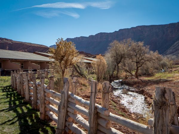 A rustic wooden fence winds beside a creek, with dry trees, a building to the left, and mountains in the background under a clear blue sky.