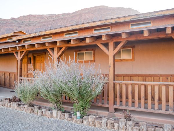 A rustic building with wooden beams, plants in front, gravel path, and a mountain backdrop under a clear sky.