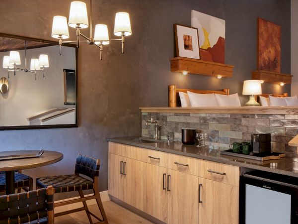 This image shows a cozy room with modern kitchen, a round table with two chairs, wall art, and a large mirror reflecting a chandelier.