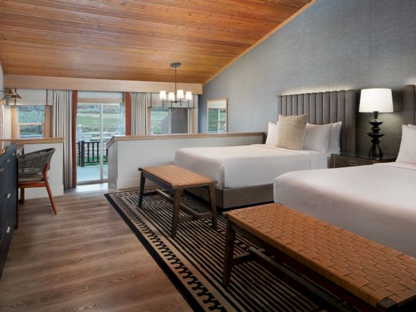A modern hotel room with two queen beds, a wooden ceiling, a desk area, neutral decor, and a balcony view.