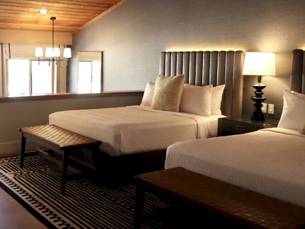 The image shows a cozy hotel room with two beds, a lamp, cushions, and a stylish rug on the floor, enhancing the inviting atmosphere.