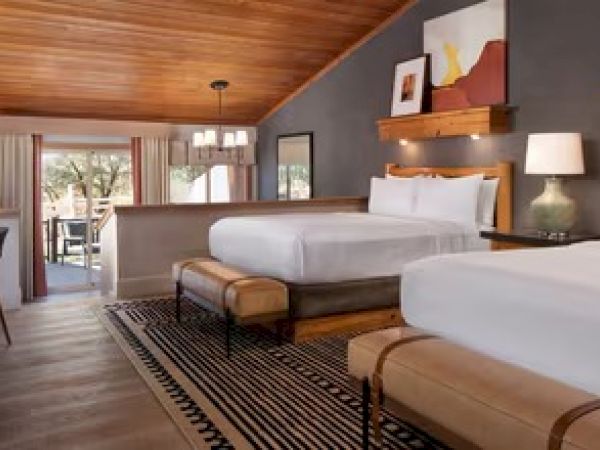 A cozy bedroom with two beds, wooden ceiling, artwork, lamps, and an outdoor view through the open doors on the left.