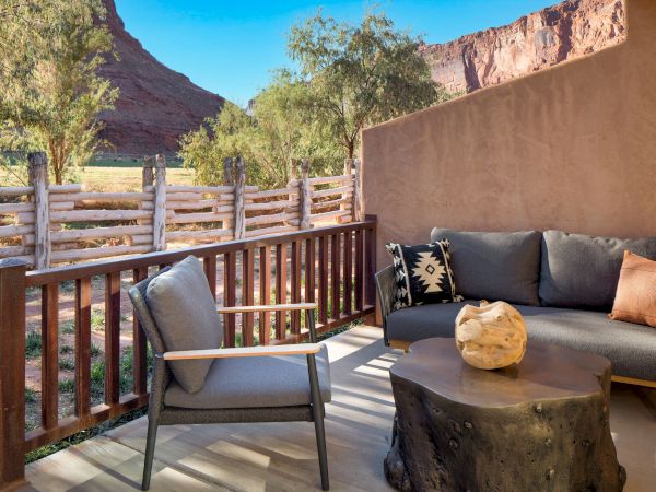 Outdoor patio with seating, a unique table, and a scenic view of a wooden fence, trees, and red rock formations under a clear blue sky.