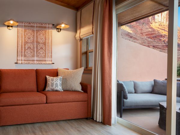A cozy room with an orange couch, wall art, and pillows opens onto a patio with outdoor seating and a view of rocky terrain.