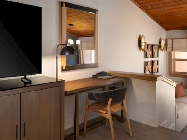 The image shows a cozy room with a TV, wooden desk, chair, mirror, wall art, and a small couch area beneath a wooden ceiling.
