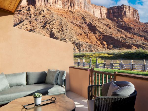 A patio with comfortable seating overlooks a river, wooden fence, and stunning red rock cliffs under a blue sky.