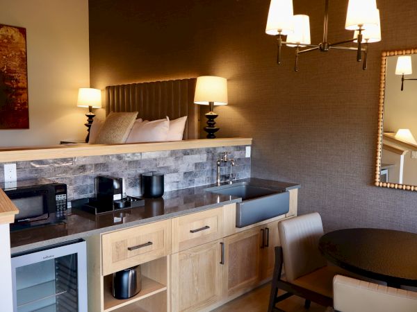 The image shows a compact kitchen area with a microwave, mini-fridge, and sink. There's a dining table, chairs, and modern decor.