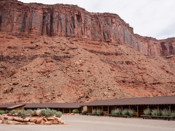 The image shows a long, low building at the base of a large red rock cliff, with a few plants in the foreground.