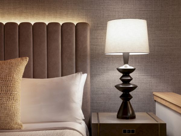 A cozy bedroom scene with a tufted headboard, white and beige pillows, a glass-top nightstand, and a modern black lamp on the side.