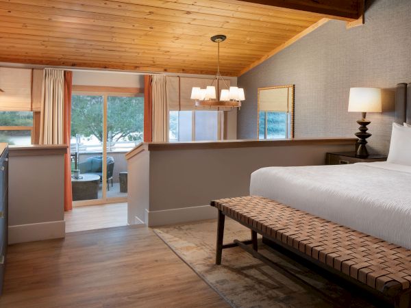 A cozy bedroom with a wooden ceiling, bed, bench, dresser, and sliding doors to a balcony with outdoor seating and a scenic view.
