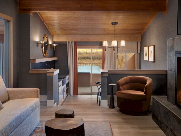 Cozy rustic living space with a sofa, round chair, fireplace, and a small dining bar; warm wood tones, soft lighting, and a lake view.