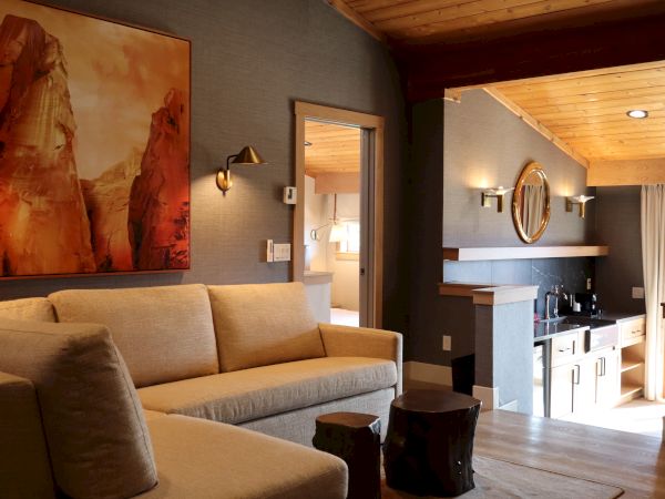 A cozy living room with a beige sofa and rustic decor, featuring a mountain painting and warm lighting from wall sconces and ceiling lights.