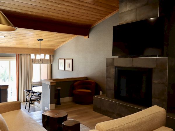 A cozy living room with a fireplace, wall-mounted TV, beige sofas, wooden ceiling, and a dining area near large windows with curtains.