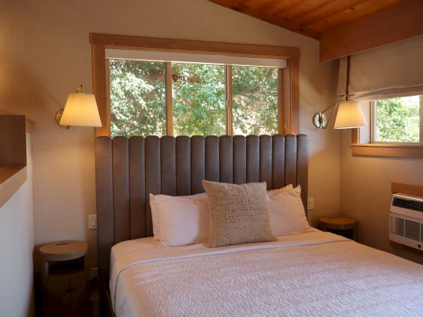 The image shows a cozy bedroom with a large bed, lamps, and windows offering a view of greenery outside.