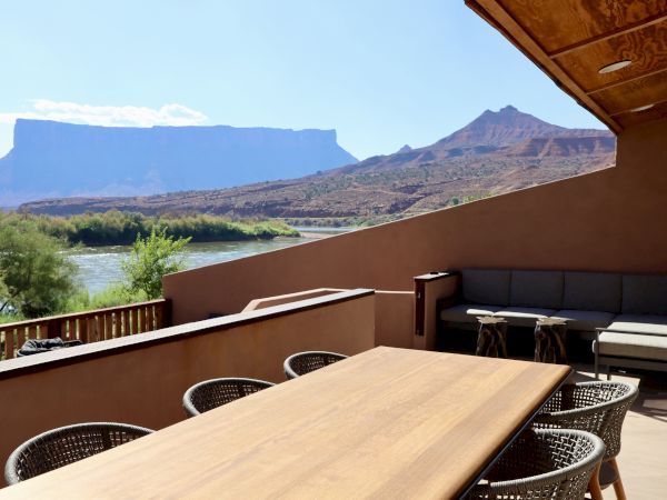 A patio with a wooden dining table and chairs overlooks a river and mountainous landscape in the background under a clear sky.