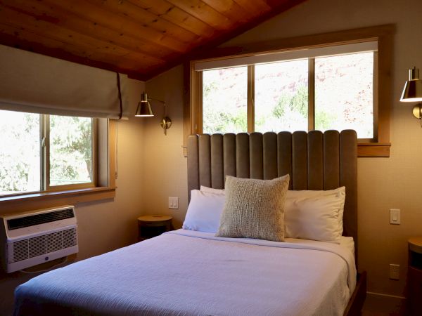 A cozy bedroom with a bed, pillows, a large window, wooden ceiling, wall lamps, and an air conditioner on the side wall.