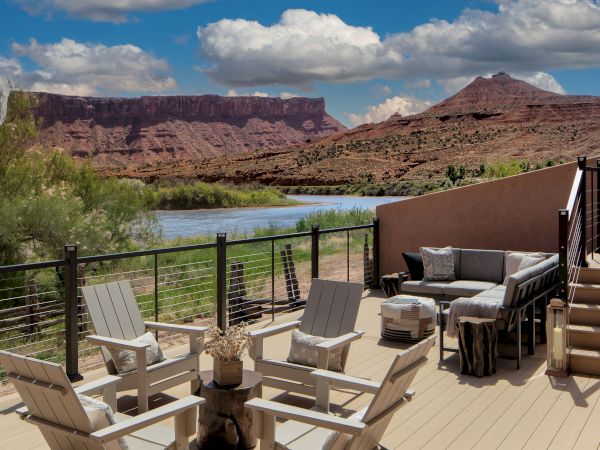Desert canyon view with a river, patio seating, and Adirondack chairs on a deck overlooking red rock mesas at sunset, inviting relaxation.