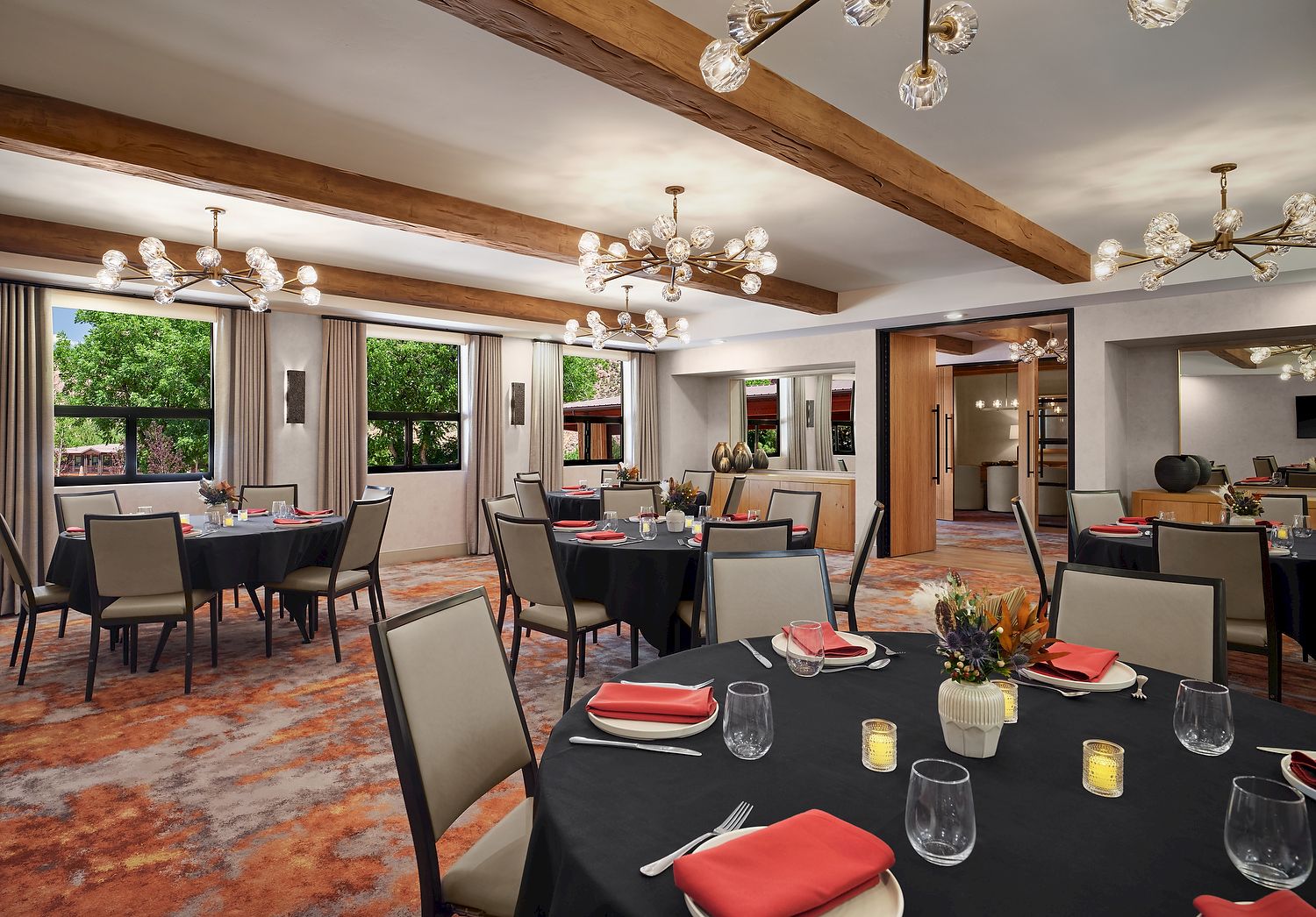The image shows a dining area with tables set with red napkins and candles, surrounded by chairs, under modern chandeliers.