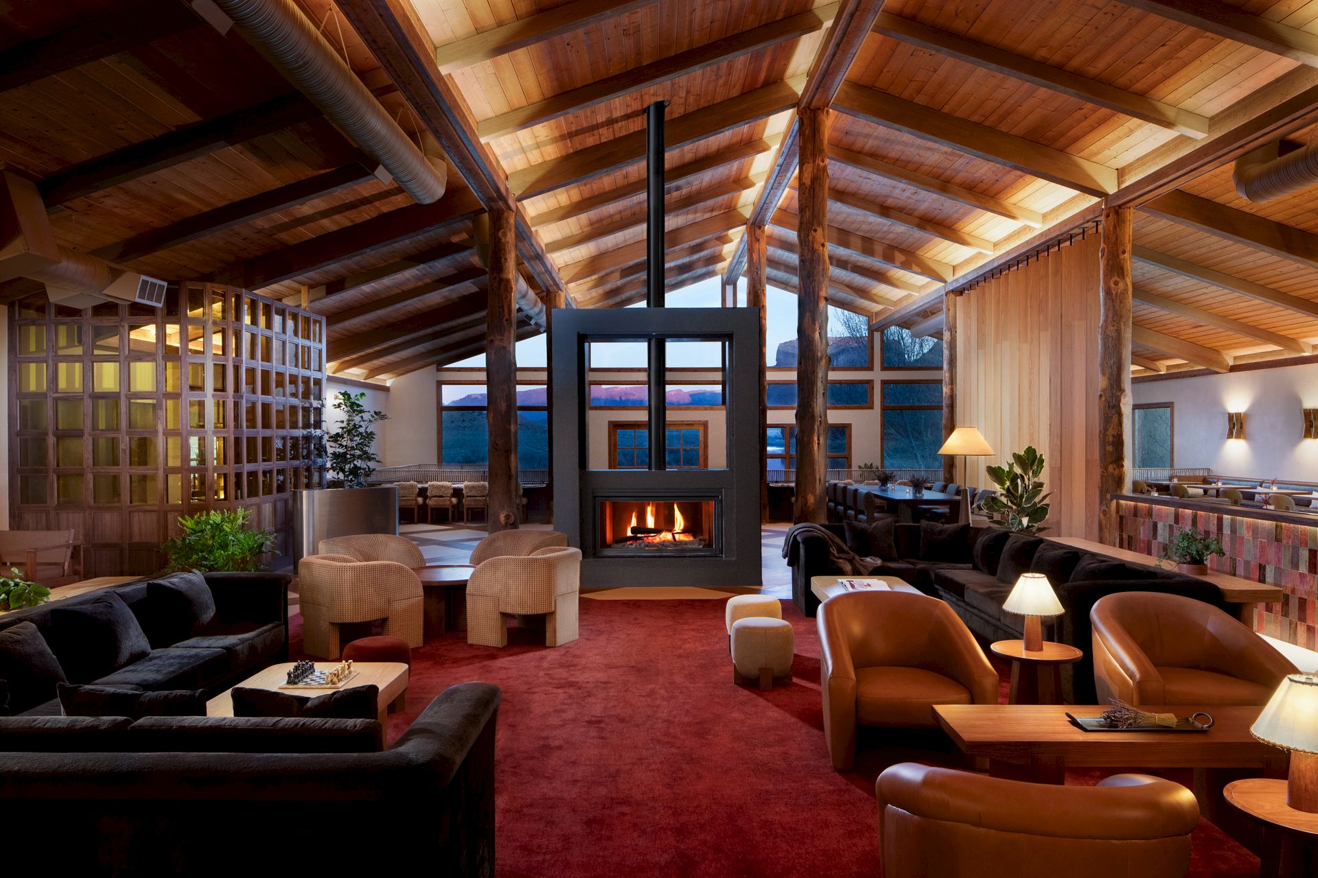 A spacious, cozy lodge-like living room with a stone fireplace, high wooden beams, red carpet, and warm seating clusters near soft lighting.