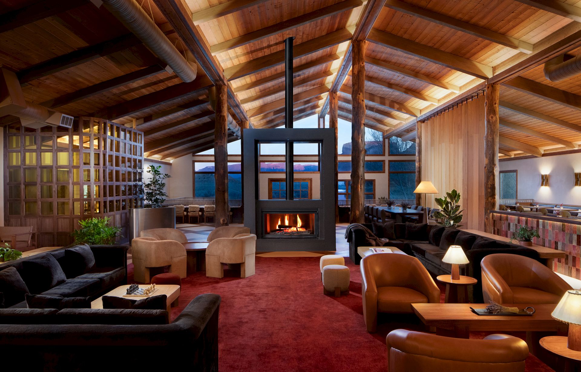 A spacious, cozy lodge-like living room with a stone fireplace, high wooden beams, red carpet, and warm seating clusters near soft lighting.