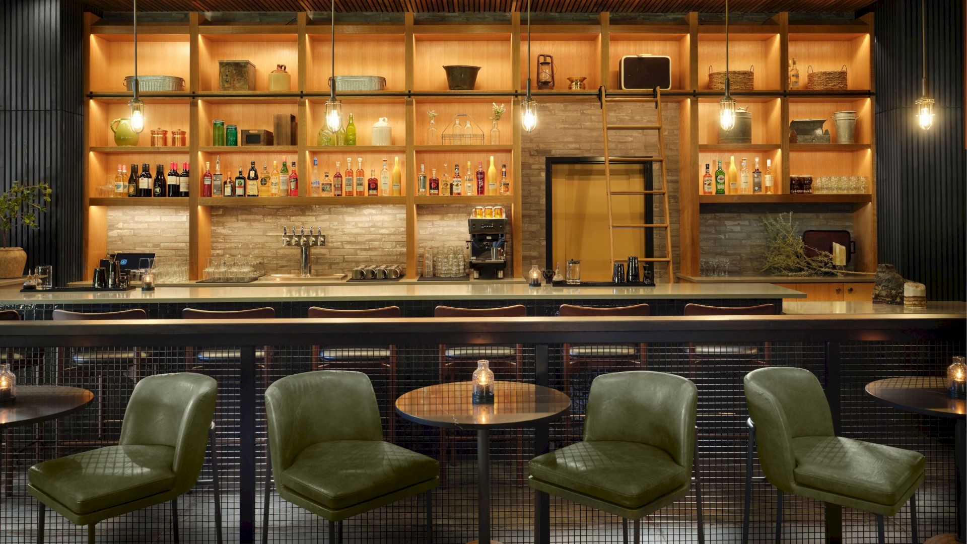 A stylish bar with dim lighting, featuring green chairs and a variety of bottles displayed on wooden shelves behind the counter.