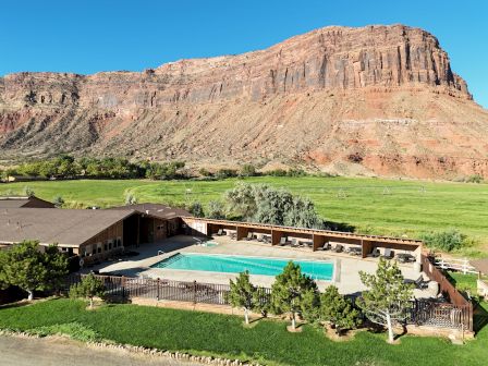 A desert resort with a rectangular pool, brown buildings, green lawns, and a towering rocky mesa in the background, under a clear blue sky.