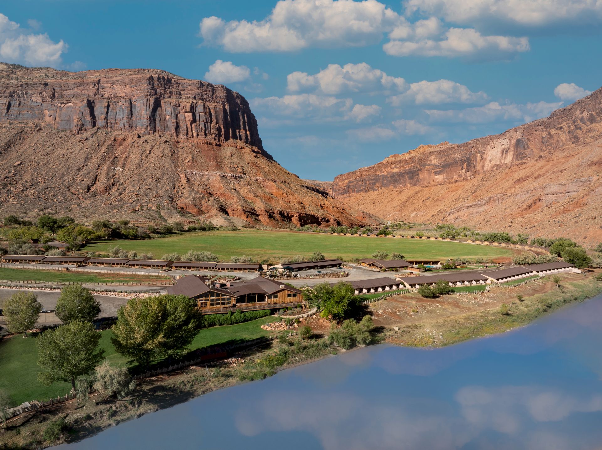 A desert canyon with towering cliffs and a riverside ranch complex, green lawns, and cabins under a blue sky with clouds.
