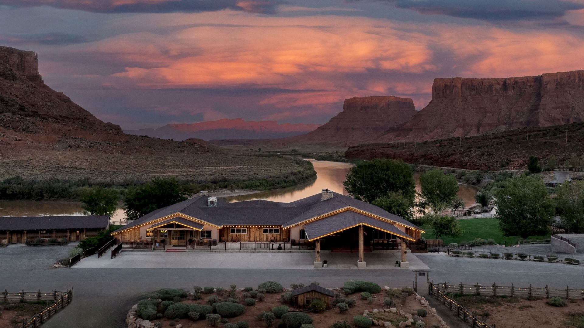 A riverside lodge with warm string lights sits beside a winding canyon river at sunset under dramatic clouds.