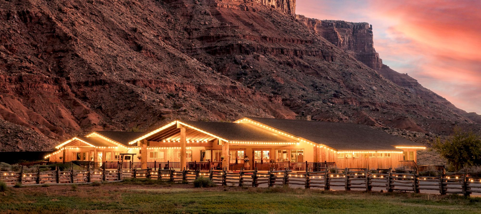 A scenic view of a lit building beneath cliffs at sunset, with a vibrant sky and natural surroundings.