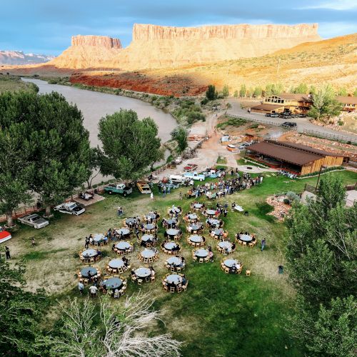 An outdoor gathering with tables and chairs on a grassy area, surrounded by trees, near a river and cliffs in the background.