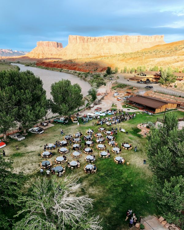 An outdoor gathering with tables and chairs on a grassy area, surrounded by trees, near a river and cliffs in the background.
