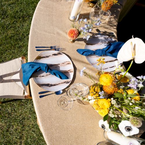 A table set for one with a circular mat, blue napkin, cutlery, and floral arrangements, on grass, next to a wooden chair.